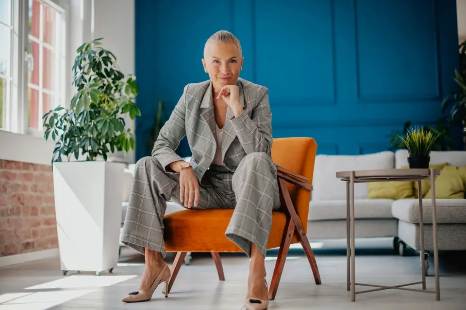 Vrouw in een grijs geruit pak, zittend op een oranje fauteuil, voor een diepblauwe wand.