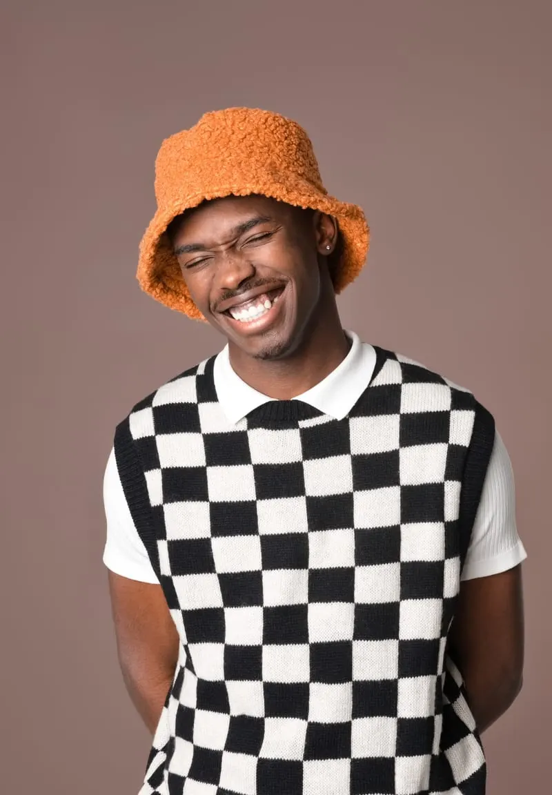 Lachende jonge man met een oranje teddy bucket hat en een zwart-wit geblokte spencer.