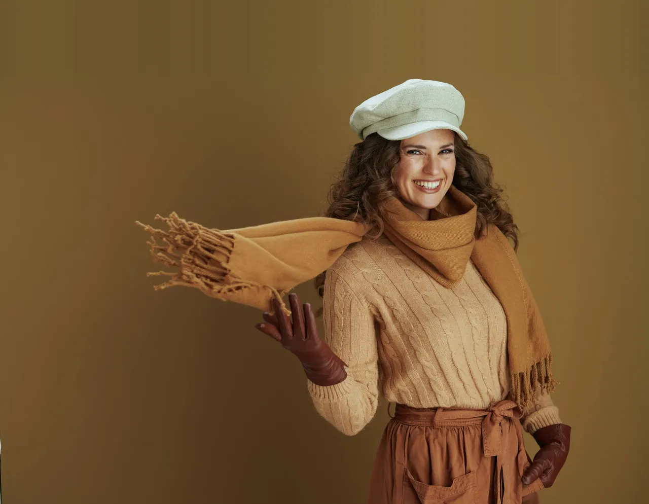 Lachende vrouw met een witte pet, een kabeltrui en een camelkleurige sjaal die ze uitzwiert, tegen een warme okerkleurige achtergrond.