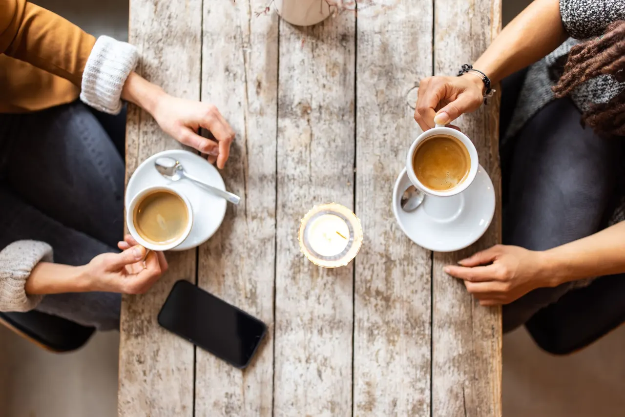 Twee mensen aan een houten tafel met koffie, van bovenaf gezien, met een kaarsje ertussen.
